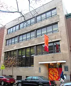 A modern-style four-story building with three rows of glass windows. The front door is painted orange with a lion motif, repeated on an orange flag hanging above the entrance.