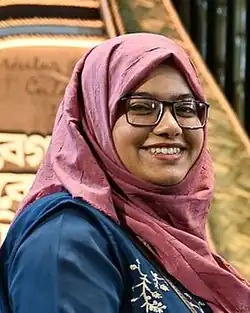 Headshot of Idris looking sideways and smiling at the camera. She wears a blue embroidered top, a pink hijab, and glasses.
