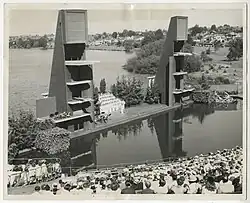 Black and white aerial photograph depicting a swimming pool surrounded by a seating area, and a stage with diving platforms on both ends