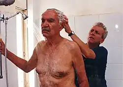 A man steadies himself by holding a grab bar in the shower, while another man shampoos his hair