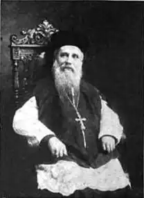 An elderly man with a long beard, a hat, and a crucifix hanging from his neck is sitting on an ornate chair.