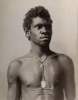 Portrait photograph of a shirtless Aboriginal man with a cord necklace and scarred torso