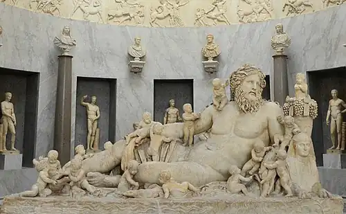A large marble sculpture, inside a formal room. The main object is a reclining nude man, surrounded by many children, as well as some animals.