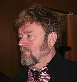 Informal, three-quarter profile, colour photo of an unsmiling man with short, curly brown hair, sideburns and a greying beard.