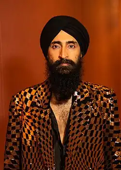Bearded man wearing a black turban and a gold-and-black sequined jacket against an orange background