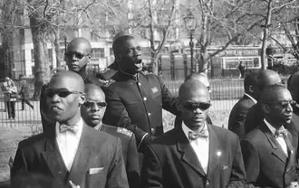 A group of young black men, wearing suits and with shaven heads, standing outdoors. One is in the center, speaking aloud. The others surround him, facing outward. Although the speaker does not wear sunglasses, the others all do.