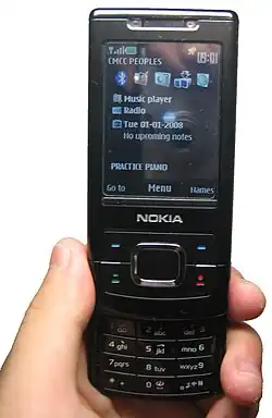 A black Nokia 6500 is held against a blank background. The phone's keypad is visible with the screen displaying the home screen.