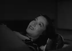 Noriko, in traditional costume, lying on a futon and covered with a blanket, stares at the ceiling, smiling