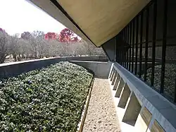 View of the exterior. The glass curtain wall is visible at right and consists of high glass panels above a sloped granite base. Part of the ceiling protrudes outward over the curtain wall, overhanging the moat to the left.