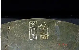 Close up of a dark stone bowl inscribed with white hieroglyphs showing a Horus falcon above a serekh and further signs on the left