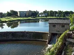 A body of water with a curved stone dam. At the right of the dam is a small stone house, underneath which water flows. There are buildings on land behind the dammed water