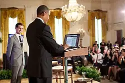 Obama in blue suit standing at a podium in front of an audience as a man in a light grey suit looks on.