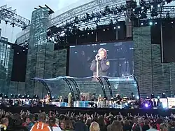 Full view of four men performing on stage which is lit by aqua-coloured lights. The most central man, dressed in mostly black, stands with his left arm reached over his head and his right arm holding out a microphone stand towards the front of the stage. A second man wearing a black tank top and maroon pants stands to the left playing his black and white guitar. Behind him is the third man with long, curly, blonde hair, suiting all black playing the keyboard. In the upper right corner is the upper body of a fourth man behind a drum kit.
