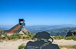 Colour photograph showing in the foreground a desman sculpted on a metal plate, and in the background a developed promontory overlooking a valley.