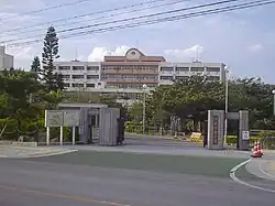 Main gate of Okinawa International University, with the gate and a road on the foreground, trees in the middle and one of the school buildings in the background