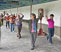 Six women, some elderly, standing in a column (except for one on the right) next to a white wall on a stone floor. All are clad in street clothes, with their left arm curled over their heads and their right arm extended.