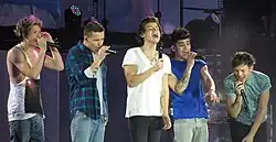 Five teenage boys standing next to each other and singing on stage. They all wear jeans, and everyone except the boy standing on the far left wears a t-shirt. The boy on the far-left wears a tank top. From left to right: Niall Horan, Liam Payne, Harry Styles, Zayn Malik, and Louis Tomlinson.