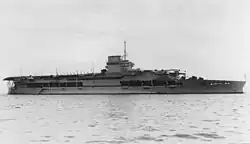 Side view of a gray anchored aircraft carrier with a short flight deck. Her island incorporates a funnel and is surmounted by a tall mast.