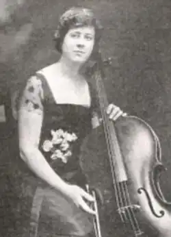 A white woman with short dark hair, wearing a dark gown with a square neckline and floral details, seated with a cello and bow