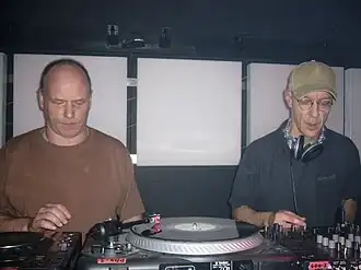 Two men perform music using various mixing boards and turntables.