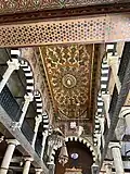 A richly decorated ceiling filled with geometric and floral patterns in warm golden tones. Arched columns in black-and-white stripes frame the hall with elegant rhythm. Hanging lamps add a sacred, timeless atmosphere to the historic synagogue interior.