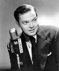 A 1941 portrait of Orson Welles dressed in a suit, speaking into a CBS microphone