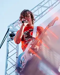 Oscar Stembridge wearing an orange Calgary Flames t-shirt, standing on an outdoor stage with an electric guitar around his neck, appearing to speak or sing into a microphone