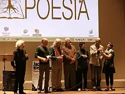 A group of smiling people on stage. There is a projected screen in the background with the word POESIA and a logo of tree growing out of a book.