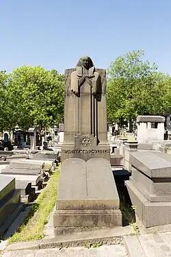 Tall stone tomb topped with carved Egyptian pharaoh figure, arms crossed, with inscriptions below, surrounded by other gravestones in cemetery