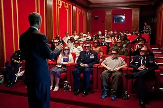 President Obama faces away from the camera towards a small movie theater filled with viewers wearing 3D glasses