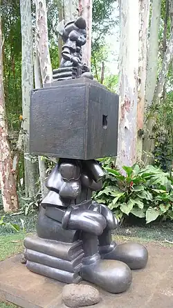 A bronze sculpture of a cartoonish figure with exaggerated limbs and shoes has a large cube for a head, with a smaller figure seated on top, set in a lush garden surrounded by tall trees