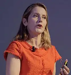 Photograph of Paola Antonelli, with a microphone and presentation clicker remote, speaking at an event