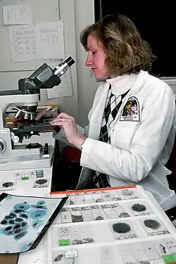 A color photo taken in 1988 shows an elegant woman manipulating sample slides on a large and fancy microscope. She is wearing a white scientific coat and is surrounded by more glass microscope slides, data sheets, and other research tools.
