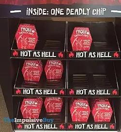 A black corrugated retail display containing red coffin-shaped boxes. The display says "Inside: One Deadly Chip" and each shelf reads "hot as hell". The shelves have about half their capacity left. The boxes are in the shape of traditional, tapered hexagonal coffins. The box says "Paqui Torilla Chips", "Carolina Reaper Madness" and has a line drawing of Death walking with a scythe.