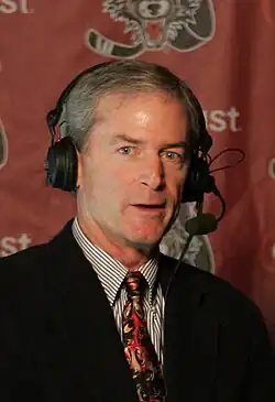 A candid bust photo of a white man with grey hair looking into the camera; he is wearing a shirt, tie, jacket, and large headset