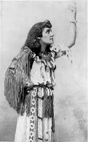 Black-and-white studio portrait of E. Pauline Johnson in profile, wearing a fringed garment and patterned sash, her hair long and loose; she raises one arm to display a bear claw bracelet on her wrist.