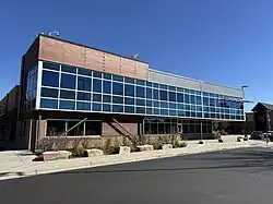 This photograph shows the contemporary counseling and classroom wing on the north side of the Peak to Peak Charter School campus in Lafayette, Colorado, featuring a two-story glass curtain wall with aluminum mullions, horizontal composite cladding above brick courses, diagonal steel braces at the base, and native plantings and stone blocks along the public sidewalk under a clear blue sky.
