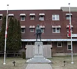 A dark bronze soldier statue stands on a light grey stone pedestal in front of a brick building. Flagpoles rise on either side, with evergreen trees and a paved plaza around the memorial.