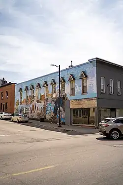 A corner building painted with a large mural dominated by blue tones, showing people and historic scenes. The artwork covers the upper wall above street-level windows, with cars parked along the street.