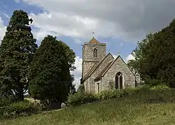The stone tower of a church seen from the east, with a pyramidal roof, standing between trees.