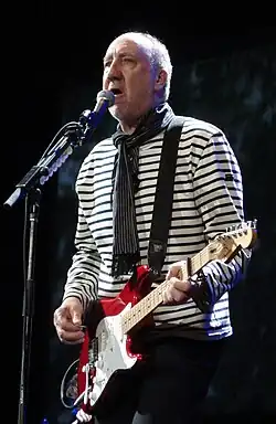 An older bald man singing into a microphone holding a red guitar.