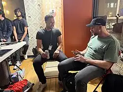 Two men sitting in folding chairs in a backstage area. One man wears a green shirt and a black baseball cap; the other wears a black t-shirt and holds a silver can. Wallpaper with various faces is visible in the background.
