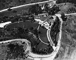 Aerial black-and-white photo showing a grand mansion with a curved driveway and manicured lawn nestled in the hills, surrounded by sparse trees and dirt roads