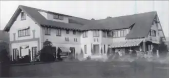 Black-and-white photo of a Tudor-style mansion with steep gabled roofs, striped awnings over the windows, and a wide lawn