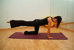 A dark-haired female instructor wearing workout clothes (a vest and flared trousers) demonstrating the pilates move "bird dog" which is performed staring on "all fours" the with opposite legs and arms outstretched