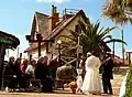 A group of seated visitors watches two actors dressed in old-fashioned nautical costumes performing outside a whimsical, red-trimmed wooden house with palm trees nearby