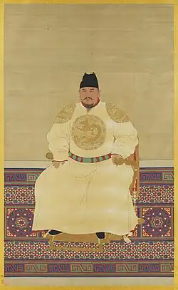 A formal, full-length portrait painting of the Hongwu Emperor seated on a throne. He wears a light yellow imperial robe embroidered with golden dragon roundels and a simple black official cap, set against a colorful patterned floor or screen in the foreground.