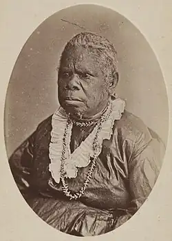 Oval-framed studio portrait of an Aboriginal woman seated, wearing a dark dress, layered necklaces, and a ruffled collar, facing slightly to the left against a plain background.