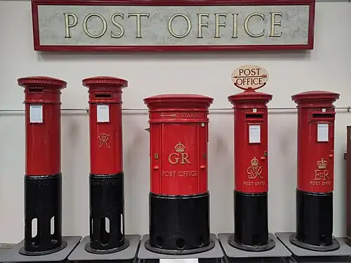 A line of 5 Red pillar boxes under a large "Post Office" sign, showing the black carcasses and how they extend below ground level