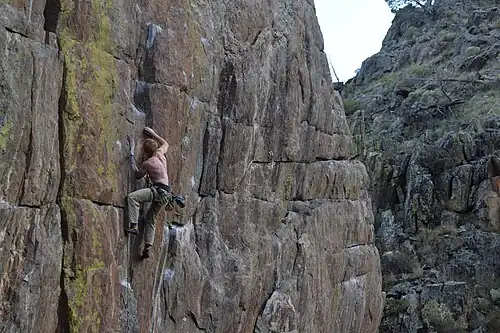Shirtless man on a steep rock face, tied on a rope, reaching up to a chalked hold with his right hand, with his left leg up.